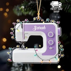 May include: A white and purple sewing machine ornament with the name "Sarah" in cursive. The ornament is decorated with colorful Christmas lights and a gold string for hanging.