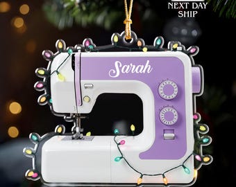 May include: A white and purple sewing machine ornament with the name "Sarah" in cursive. The ornament is decorated with colorful Christmas lights and a gold string for hanging.