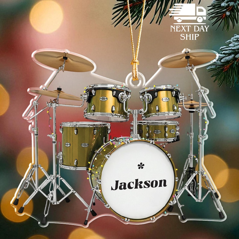 May include: A clear acrylic drum set ornament with gold-colored drums and cymbals. The bass drum has the name "Jackson" printed on it. The ornament is strung with a gold cord and has a "Next Day Ship" graphic.
