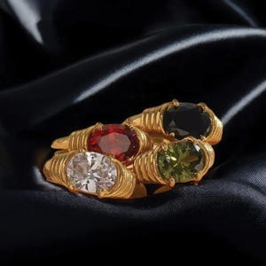 May include: Four gold-colored rings with oval-shaped gemstones in red, black, green, and white. The rings have a textured band design and are displayed on a dark, silky fabric.