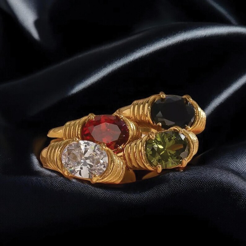 May include: Four gold-colored rings with oval-shaped gemstones in red, black, green, and white. The rings have a textured band design and are displayed on a dark, silky fabric.