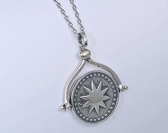 May include: A silver pendant necklace featuring a circular charm with a starburst design. The charm is suspended from a delicate chain. The pendant has a rotating design and is made of a shiny, metallic material.
