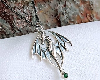 May include: A silver dragon pendant with green gemstone eyes, hanging from a black chain. The dragon is detailed with wings and a tail.