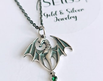 May include: A silver dragon pendant necklace with a green gemstone. The dragon is detailed with wings and a long tail. The necklace is on a black chain. The pendant is from Selliss Gold & Silver Jewelry.