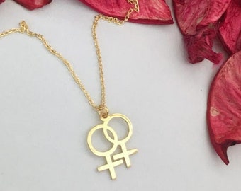 May include: A gold necklace with a double female symbol pendant. The necklace is lying on a white surface with dried red rose petals.