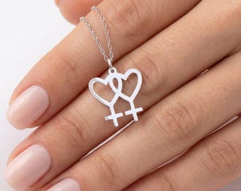 May include: A silver necklace featuring a pendant with two intertwined heart-shaped symbols. The pendant is connected to a delicate chain. The necklace is displayed on a light-toned background.