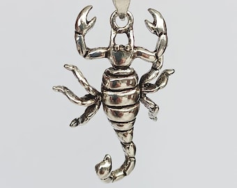 May include: A silver scorpion pendant hangs from a delicate chain. The scorpion is detailed with claws, legs, and a curved tail. The pendant is made of a shiny, metallic material and is set against a plain background.