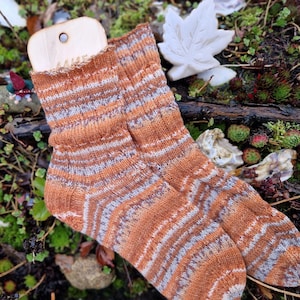 Könnte beinhalten: Ein Paar handgestrickte Socken mit orangefarbenen, grauen und weißen Streifen. Sie werden mit einem hölzernen Sockenformer präsentiert. Das Wort "AALIE" ist auf dem Former zu sehen. Die Socken liegen auf Moos und Blättern.