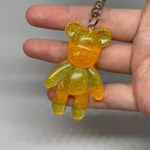 May include: A glittery, yellow and orange teddy bear keyring. The bear is made of translucent resin and has a silver metal keyring attached. The bear's body is a gradient of yellow to orange, with glitter embedded throughout.