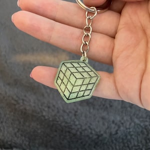May include: A green and black keychain featuring a 3D cube design. The cube is made of a translucent material with black lines outlining the cube's shape.