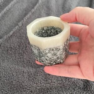 May include: A small, hexagonal container with a cream-coloured top and a textured, silver-coloured base. The inside is filled with small, dark grey, shiny pieces. The container is held in a hand against a grey, textured background.