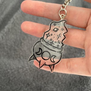 May include: A clear acrylic keychain with a celestial design. The keychain features a black outline of a rocket ship shape with a planet, stars, and crescent moons. The keychain is attached to a silver metal keyring.