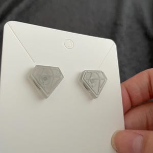 Puede incluir: Un par de pendientes en forma de diamante de color gris claro. Los pendientes se muestran en una tarjeta blanca. Los pendientes tienen un diseño geométrico dentro de la forma de diamante.