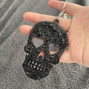 Puede incluir: Llavero negro con forma de calavera con recortes en forma de corazón para los ojos y una mandíbula dentada. El llavero está cubierto de purpurina negra brillante y sujeto a una cadena plateada. Un accesorio único.