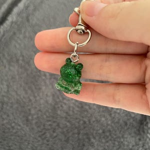 Puede incluir: Un pequeño dije de rana verde con purpurina incrustada en la resina. La rana está unida a un cierre y un anillo de metal plateado. El dije se sostiene en la mano sobre un fondo gris.