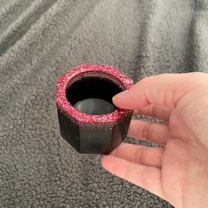 以下が含まれることがあります： 幾何学的な形状の黒い容器で、ピンク色のキラキラした縁取りが施されています。容器は手で持たれており、そのデザインと質感を際立たせています。内側は暗く、明るい縁取りと対照的です。小さな植物や装飾品に適しています。