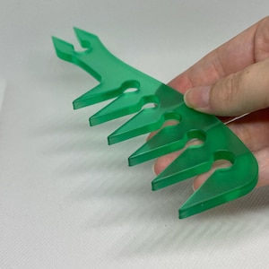 May include: A translucent green hair comb featuring a series of wide, pointed teeth with circular cutouts at the base of each gap. The comb has a smooth, flat surface and a distinct, angular design with five prominent teeth for detangling hair.