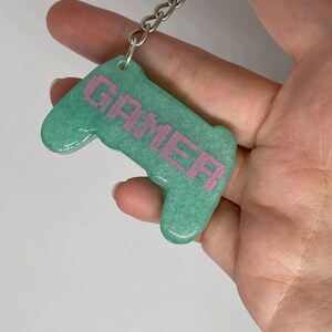 May include: A teal-colored keychain in the shape of a video game controller. The word "GAMER" is printed in pink pixelated letters across the controller. The keychain is attached to a silver chain and ring.