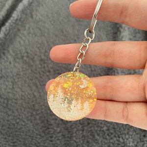 Puede incluir: Un llavero redondo con una escena de bosque. El llavero tiene una cadena y un anillo plateados. La escena del bosque presenta árboles blancos, una luna creciente y purpurina amarilla y verde sobre un fondo naranja.
