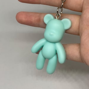 以下が含まれることがあります： ミントグリーンのクマの形をしたチャームで、シルバーカラーの金属製キーホルダーに取り付けられています。クマのフィギュアは滑らかなマット仕上げで、丸い耳、シンプルな顔の構造、関節のある手足が特徴で、金属製のリンクチェーンから吊り下げられています。