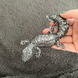 May include: A silver glitter gecko figurine with a curled tail. The gecko is posed with its legs outstretched and its head tilted up.