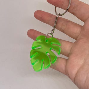 May include: A neon green Monstera leaf keychain. The leaf-shaped charm is attached to a silver chain and keyring. The keychain is made of a translucent, bright green material.