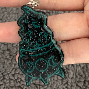May include: Black and green glitter resin keychain shaped like a cauldron with a crescent moon and stars design.