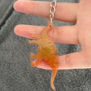 May include: A gold and orange dinosaur keychain. The dinosaur is a stylized silhouette with a textured, glittery appearance. The keychain has a silver chain and ring for attachment.