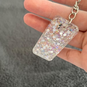 Puede incluir: Un llavero plateado con un dije rectangular lleno de purpurina iridiscente. El dije es transparente y tiene una variedad de colores, incluyendo rosa, blanco y plateado.