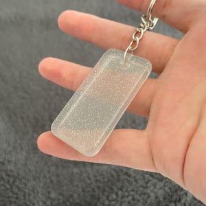 May include: A clear, rectangular keychain with silver glitter embedded in the plastic. The keychain has a silver metal ring attached to it.