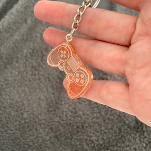 May include: Orange and silver glitter resin keychain shaped like a video game controller. The keychain has a silver chain and key ring. The controller design includes buttons and a directional pad.