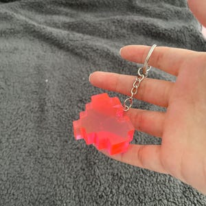 May include: A neon pink pixelated keyring with a silver chain. The keyring is held against a grey, textured background. The pixelated design gives the keyring a retro, video game aesthetic.