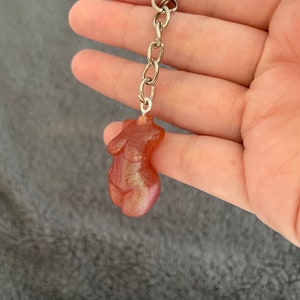 May include: A silver keychain with a red-brown resin figurine of a female torso. The figurine has a glittery finish.