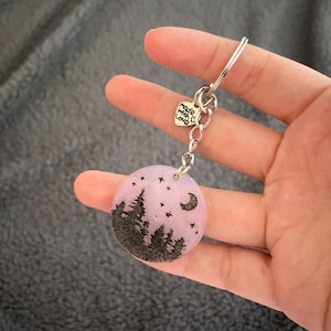 May include: A circular keyring with a silver-tone chain and ring. The pendant is a pale purple colour, featuring a black forest silhouette, stars, and a crescent moon. A small heart-shaped charm reads "made with love."