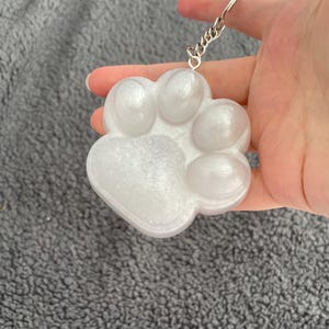 Puede incluir: Un llavero blanco nacarado con forma de huella de pata con una cadena plateada. La huella está hecha de un material transparente y brillante. El llavero se sostiene en una mano sobre un fondo gris.