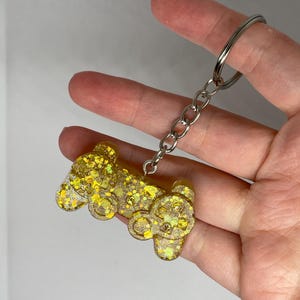 May include: A glittery gold keychain featuring a bear design. The bear is made of clear resin filled with gold glitter and has a silver chain and keyring. The keychain is held in a hand.