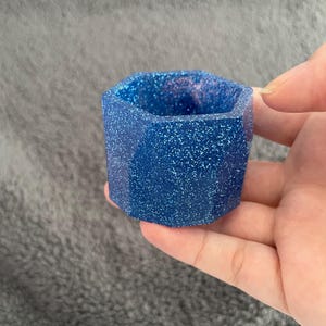 Puede incluir: Un pequeño macetero hexagonal hecho de resina azul con purpurina. El macetero mide aproximadamente 5 cm de alto y 5 cm de ancho.