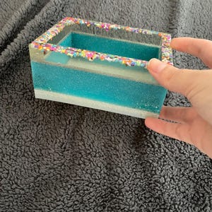 Puede incluir: Una caja rectangular de resina con interior turquesa y exterior transparente. El borde superior está decorado con chispas de colores. La caja está dividida en dos compartimentos y es sostenida por una mano.