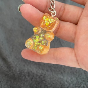 May include: A clear, gummy bear-shaped keyring with gold and iridescent glitter suspended within. The keyring is attached to a silver metal ring and chain. The bear is held in a hand against a grey background.
