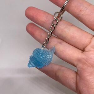 May include: A keychain featuring a translucent, light blue, textured seashell charm attached to a silver-toned metal split ring by a short chain. The seashell charm has a bumpy, crystalline surface that catches the light, creating a sparkling effect.