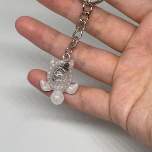 Puede incluir: Un llavero de resina transparente con forma de tortuga y una pequeña concha dentro. La tortuga tiene una cadena plateada y un llavero. El llavero se sostiene en una mano.