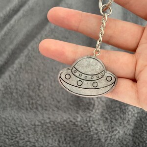 May include: A silver keyring with a small, grey, UFO-shaped charm. The charm has a detailed outline with circular windows and a domed top. The keyring includes a silver chain and a split ring for attaching keys.
