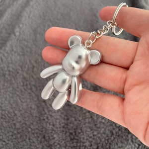 May include: A silver bear keyring. The bear-shaped charm has a smooth, reflective finish and is attached to a chain and a keyring. The bear's arms and legs are extended, and it has small, rounded ears.