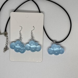 May include: A light blue cloud-shaped jewelry set, including a necklace and a pair of earrings. The necklace has a black cord, and the earrings have silver hooks. The jewelry is displayed on a white card.