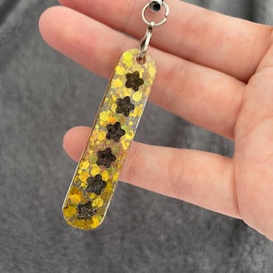 Puede incluir: Un dije rectangular y brillante de color amarillo con formas de flores negras. El dije está sujeto a un cordón negro con un cierre de metal plateado. El dije se sostiene en la mano de una persona.
