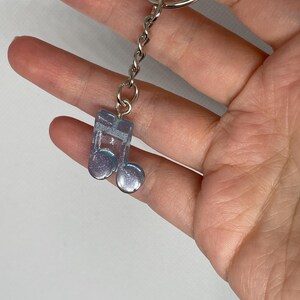 Puede incluir: Un llavero transparente e iridiscente con forma de nota musical, con una cadena y un llavero plateados. La nota musical tiene un efecto brillante y nacarado, con una combinación de colores azul claro y morado. Accesorio decorativo.