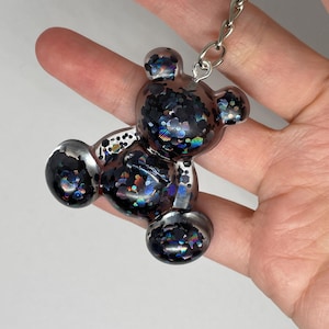 May include: A clear resin bear-shaped keyring with silver detailing and filled with iridescent glitter. The keyring is attached to a silver chain and ring.