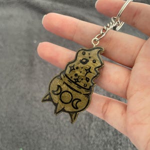 Puede incluir: Un llavero de color dorado con una ilustración en blanco y negro de un caldero con una luna creciente y estrellas. El llavero está unido a una cadena de metal plateado.
