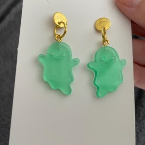 May include: A pair of earrings featuring a gold-colored stud and a green ghost-shaped charm. The ghost charms have a smiling face and are suspended from the gold studs with a small gold loop. The earrings are displayed on a white card.