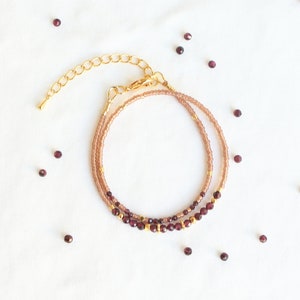 May include: A delicate gold chain bracelet with two rows of small, dark red beads. The bracelet is on a white background with scattered red beads.